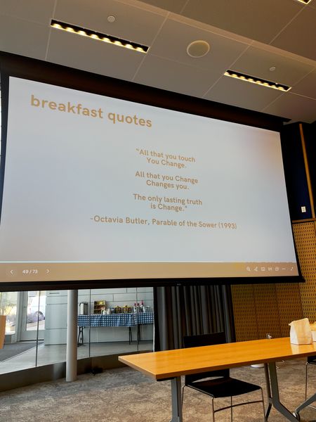 A photo of a slideshow presentation and a desk. On the presentation is a quote from Parable of the Sower by Octavia Butler: 'All that you touch / You Change. / All that you Change / Changes you. / The only lasting truth / is Change.'